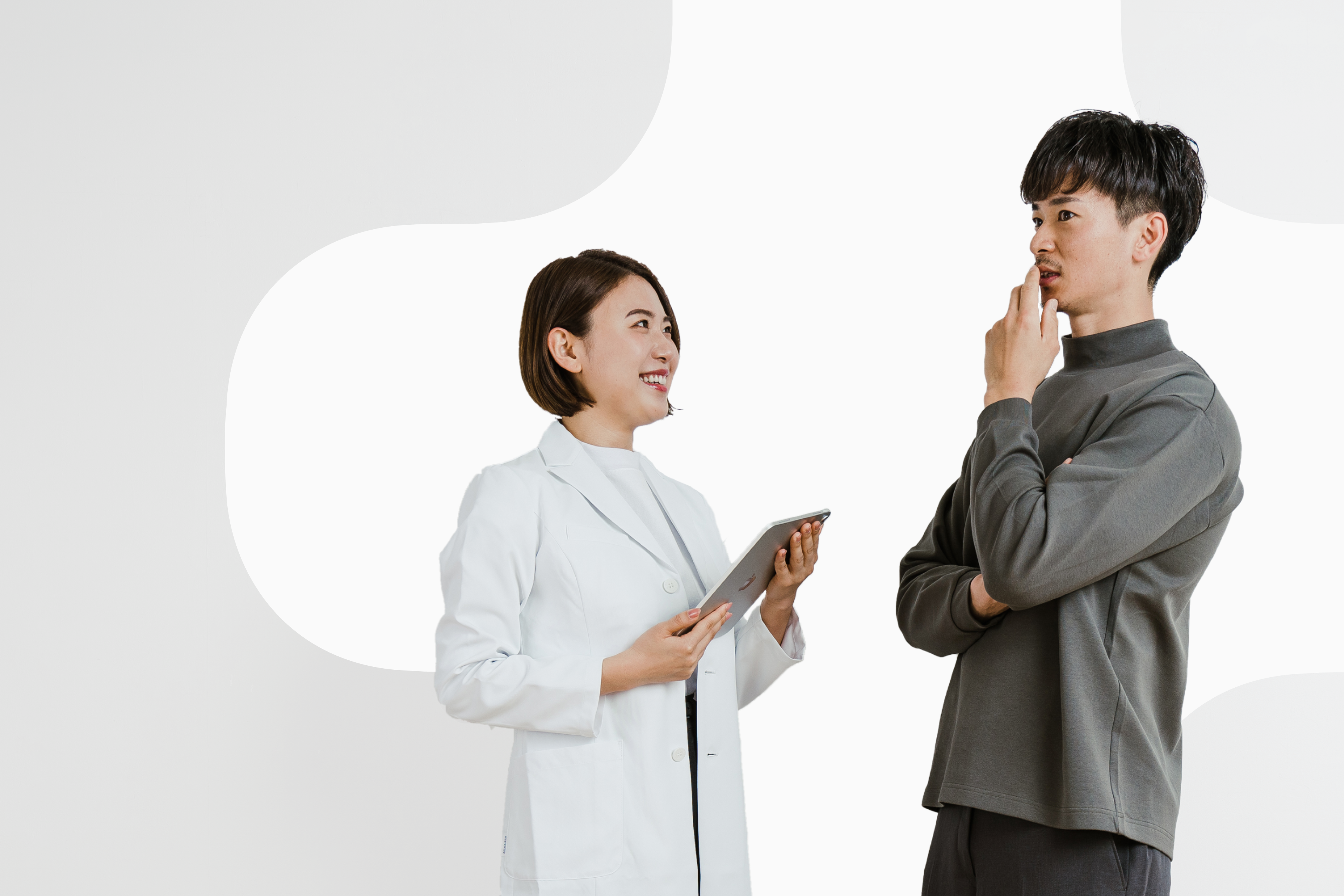 The image depicts a young man and woman. The woman is wearing a white coat and smiling. The man is wearing a gray shirt and looking at the woman with a thoughtful pose. They seem to be discussing or contemplating something. The background is completely white, making the individuals stand out. Despite the professional atmosphere, their natural expressions convey a sense of friendliness.