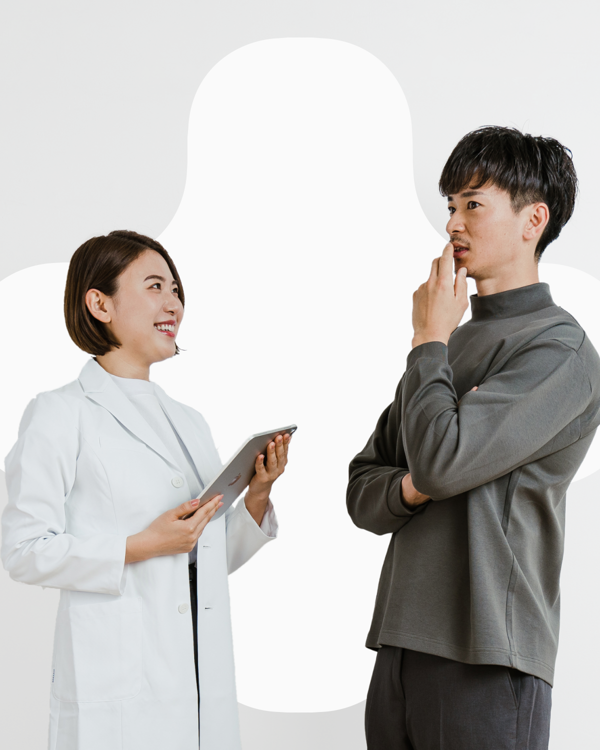 The image depicts a young man and woman. The woman is wearing a white coat and smiling. The man is wearing a gray shirt and looking at the woman with a thoughtful pose. They seem to be discussing or contemplating something. The background is completely white, making the individuals stand out. Despite the professional atmosphere, their natural expressions convey a sense of friendliness.